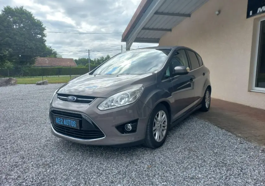 Ford C-MAX gris en 3/4 avant droit, garé sur gravier devant un bâtiment avec ciel nuageux.