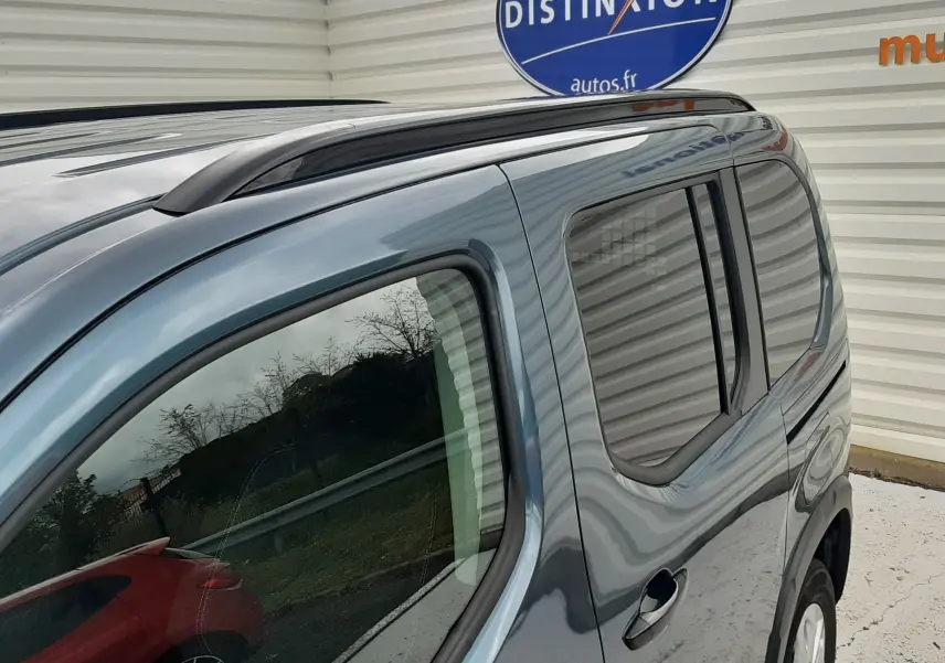 Vue 3/4 arrière côté droit du Peugeot Rifter bleu Kiama avec barres de toit noir mat et vitres arrière surteintées.