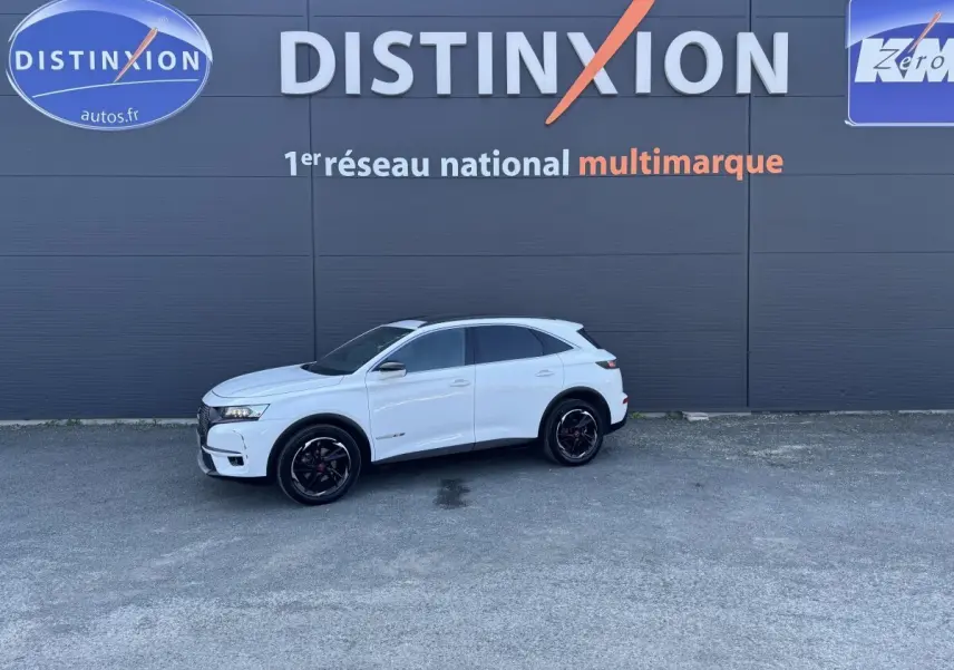 Vue de profil droit d'un DS7 Crossback blanc stationné devant un mur gris avec logo Distinxion.