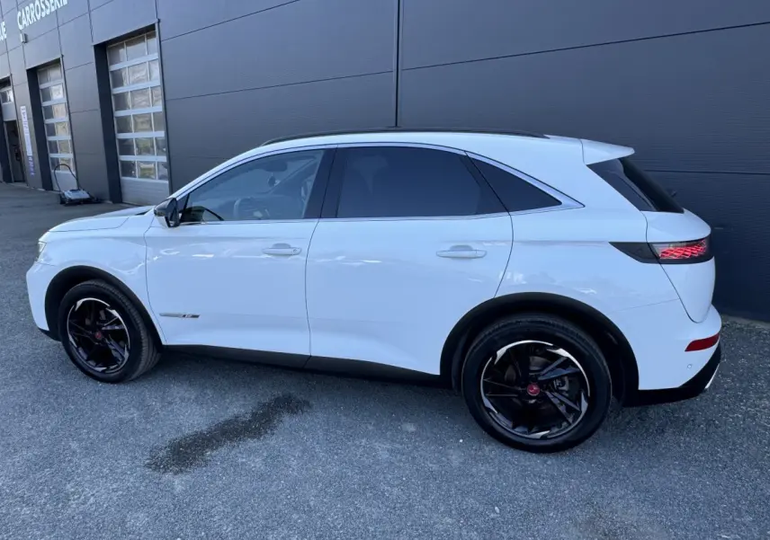 Vue de profil côté gauche d'un DS7 Crossback blanc 2020 avec jantes noires et vitres teintées, stationné devant un bâtiment.