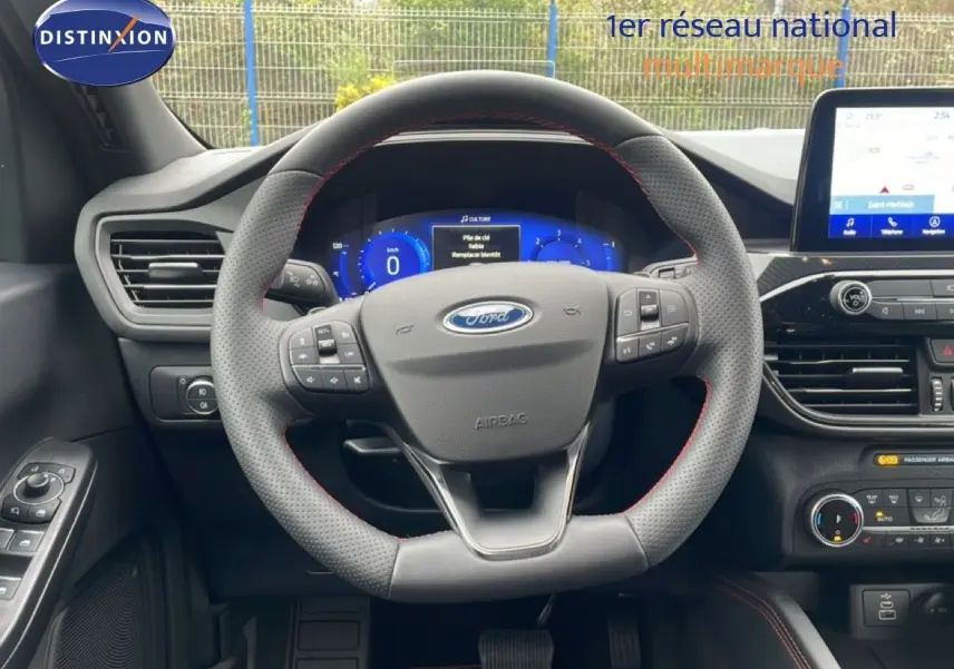 Vue intérieure centrée sur le volant perforé avec coutures rouges du Ford Kuga 2024, tableau de bord digital et écran tactile visible.