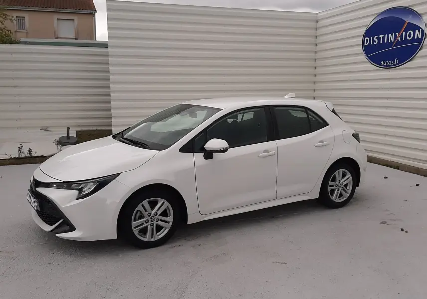 Vue de profil côté gauche d'une Toyota Corolla blanche 5 portes, hybride essence, garée devant un mur blanc avec logo Distinxion.