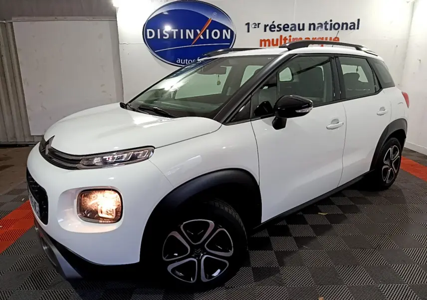 Vue 3/4 avant droite d'un Citroën C3 Aircross blanc avec phares allumés et barres de toit noires en intérieur showroom.