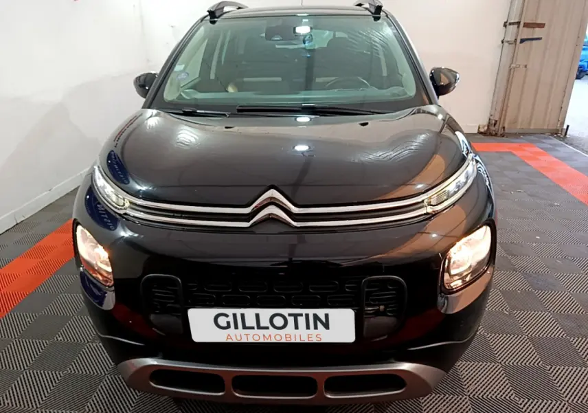 Vue avant d'un Citroën C3 Aircross noir avec phares allumés et barres de toit visibles en intérieur showroom.