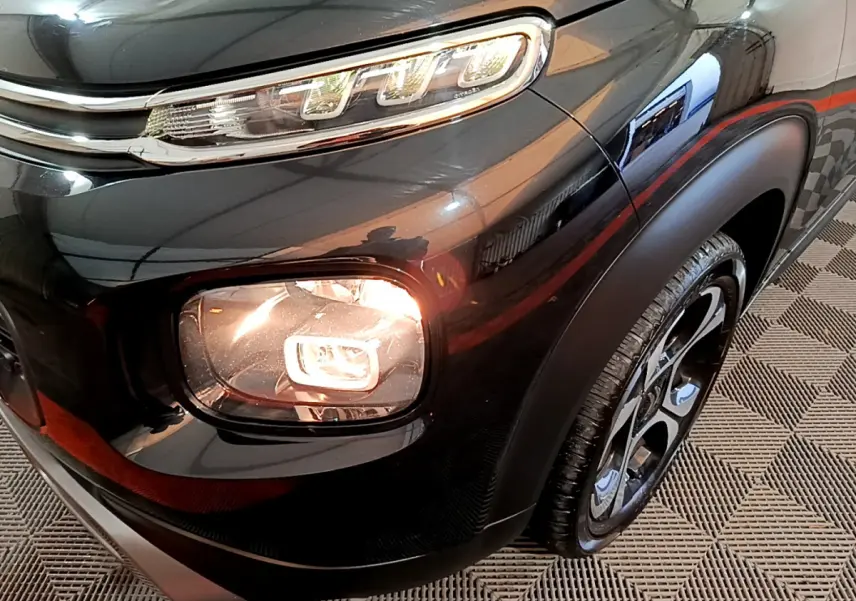 Gros plan sur le phare avant droit allumé d'un Citroën C3 Aircross noir, avec jante et passage de roue visibles.