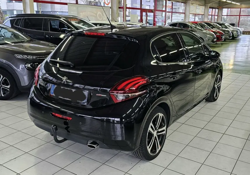Vue 3/4 arrière droite d'une Peugeot 208 GT-LINE noire avec jantes alliage et attelage dans un showroom.