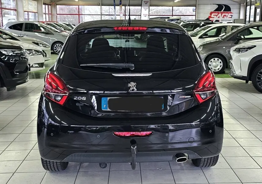 Vue arrière d'une Peugeot 208 GT-LINE noire stationnée en intérieur avec feux arrière allumés et attelage visible.