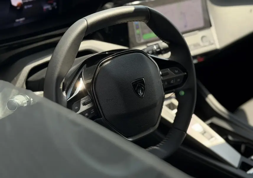Gros plan sur le volant cuir noir de la Peugeot 308 2025, avec tableau de bord numérique et écran tactile en arrière-plan.