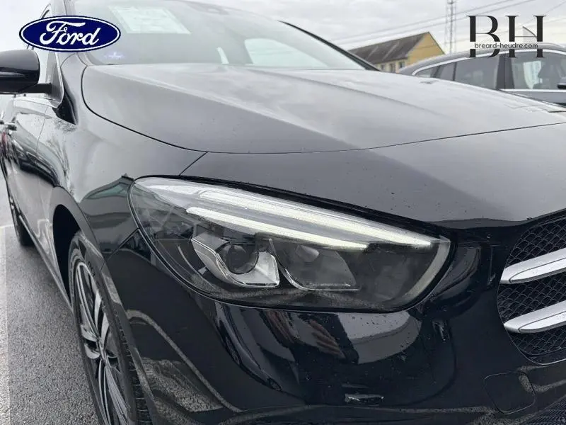 Gros plan sur le phare avant gauche d'une Mercedes Classe B noire cosmos métallisé, avec calandre et jante visibles.