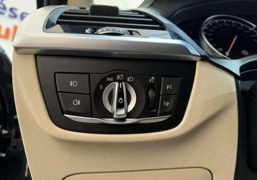 Gros plan sur les commandes d’éclairage avec molette et boutons, tableau de bord beige d’une BMW X3 gris foncé 2018.