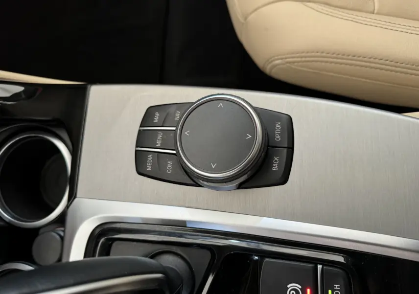 Gros plan sur la molette de commande iDrive et console en aluminium brossé, siège beige visible, BMW X3 gris foncé intérieur.