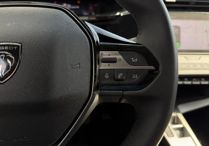 Gros plan sur le volant cuir noir de la Peugeot 308 2025 avec commandes intégrées et écran tactile en arrière-plan.