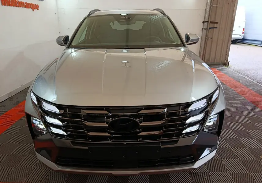 Vue avant d’un Hyundai Tucson gris clair 2025 avec calandre noire et feux LED distinctifs, en intérieur showroom.