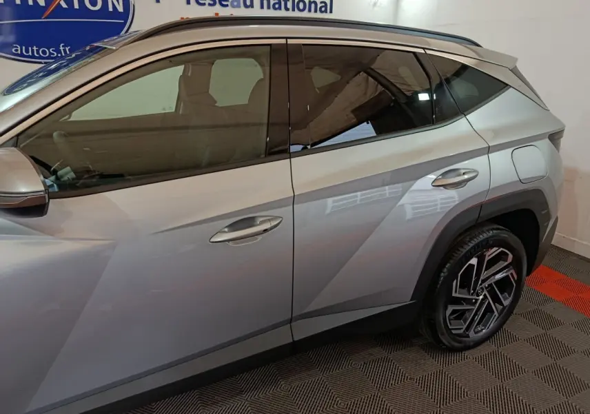 Vue latérale côté gauche d'un Hyundai Tucson 1.6 Hybrid gris clair avec jantes alliage et lignes anguleuses marquées.