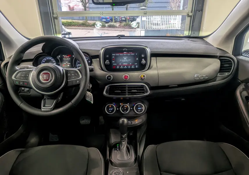 Vue intérieure frontale du tableau de bord noir du Fiat 500X 2024 avec écran tactile central et volant multifonction.