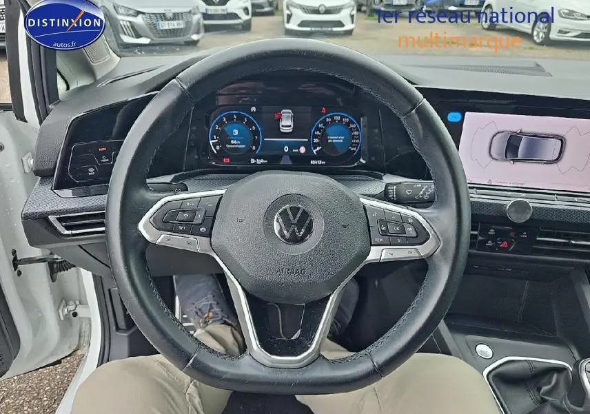 Vue intérieure centrée sur le volant cuir noir d'une Volkswagen Golf blanche, avec tableau de bord numérique et écran tactile visible.