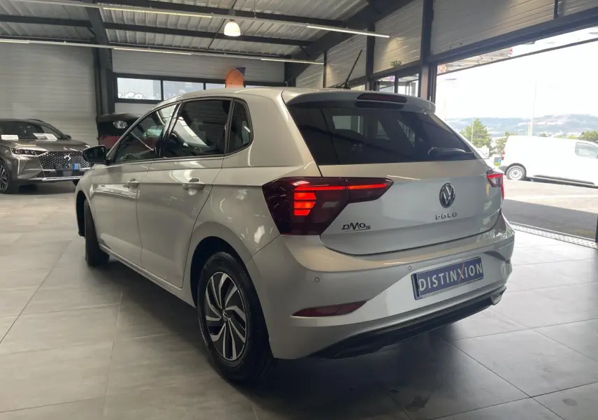 Volkswagen Polo gris clair vue 3/4 arrière droit en intérieur avec feux arrière allumés et vitres teintées.