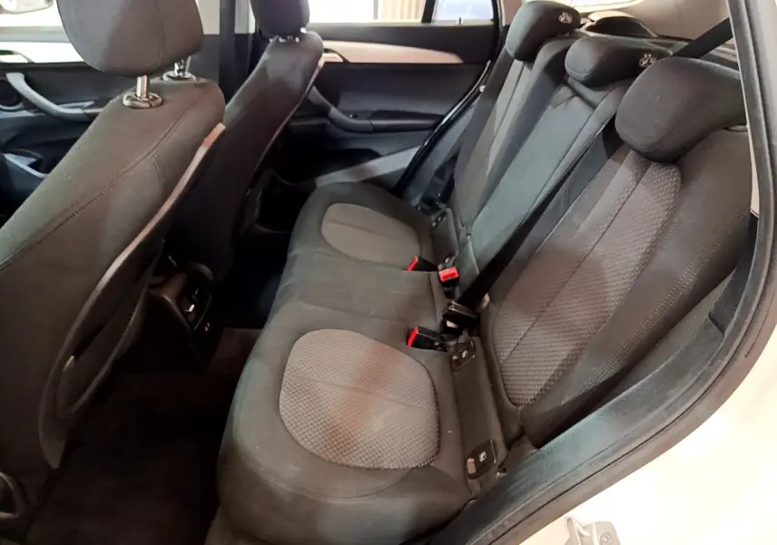 Vue intérieure arrière droite des sièges noirs du BMW X1 sDrive18i Lounge blanc de 2019, avec ceintures et appuie-têtes visibles.