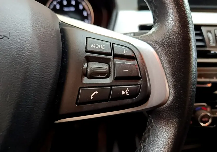 Gros plan sur les commandes gauche du volant cuir noir du BMW X1 sDrive18i Lounge 2019, avec boutons multifonctions.