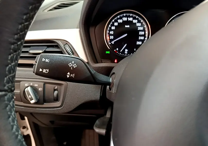 Gros plan sur le volant cuir et le compteur noir du tableau de bord d'une BMW X1 sDrive18i Lounge blanche, vue côté conducteur.