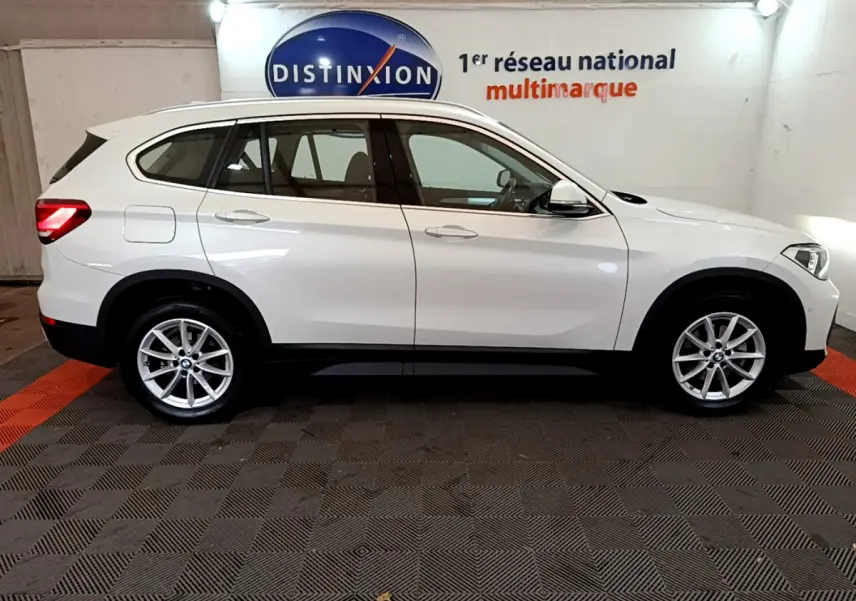 BMW X1 sDrive18i blanc vue profil côté gauche dans un showroom avec jantes alliage et barres de toit noires.