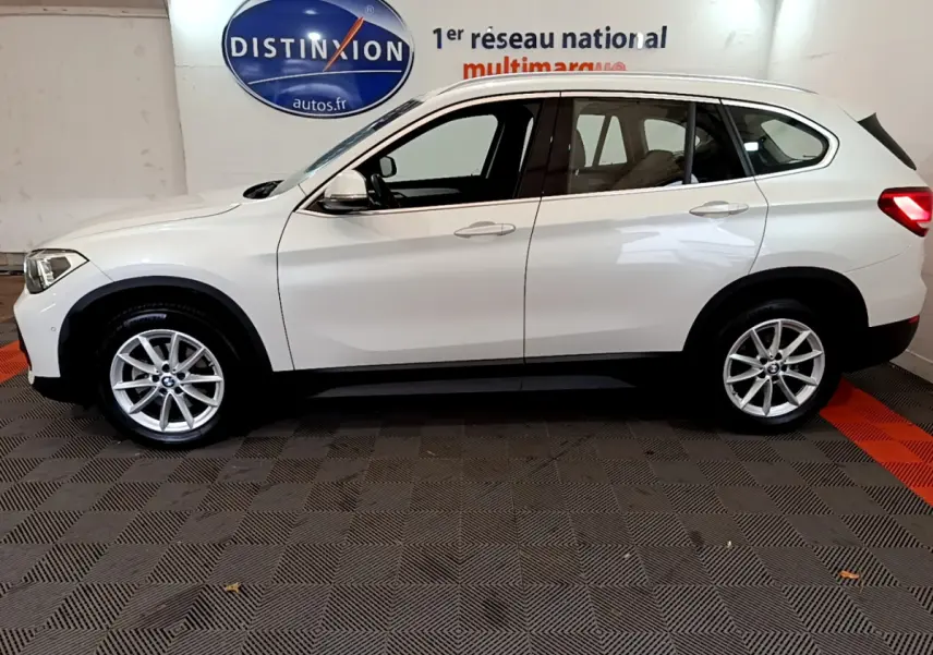 Vue de profil côté gauche d'un BMW X1 sDrive18i Lounge blanc, avec jantes alliage et rails de toit noirs.