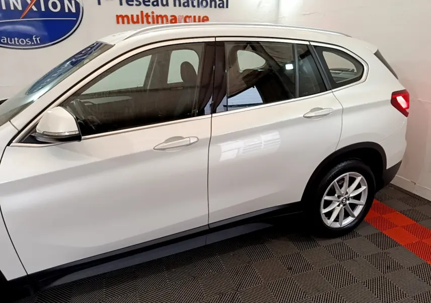 BMW X1 sDrive18i blanc vue de profil côté gauche en intérieur avec jantes alliage et rétroviseurs blancs.