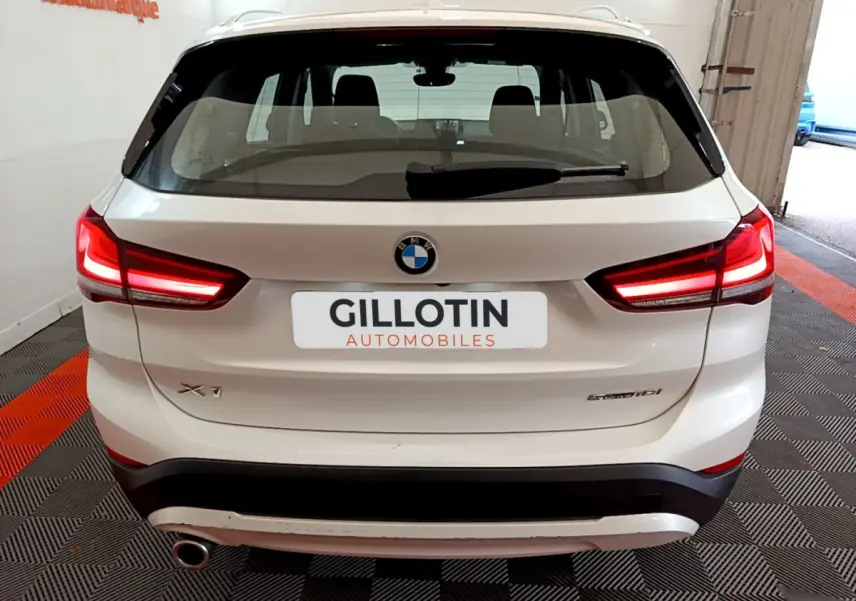 Vue arrière d'un BMW X1 sDrive18i blanc avec feux LED allumés, dans un garage au sol carrelé noir et rouge.
