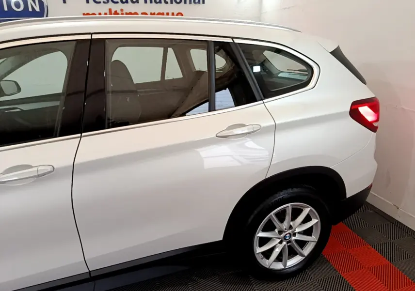 Vue côté droit arrière d'un BMW X1 sDrive18i Lounge blanc avec feux arrière allumés dans un showroom.