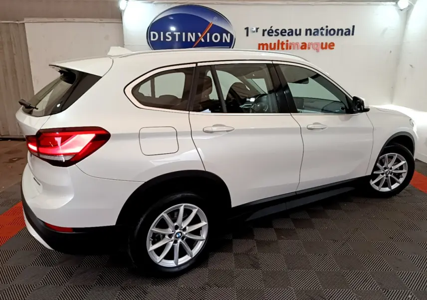 BMW X1 sDrive18i blanc en vue profil droit, feux arrière allumés, roues alliage et toit avec rails noirs.