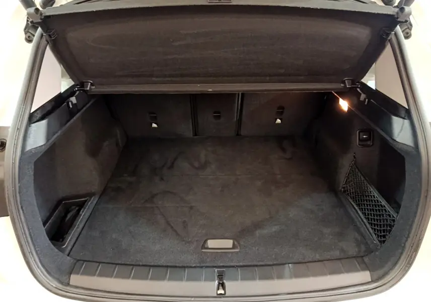 Coffre ouvert de BMW X1 sDrive18i blanc, vue arrière, espace de chargement avec tapis noir et filet latéral.