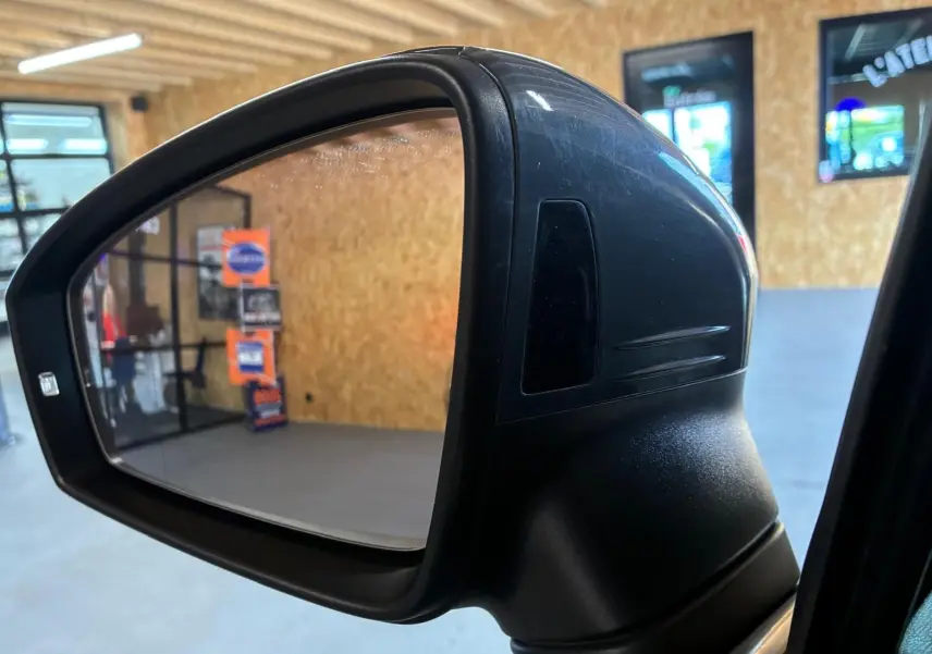 Gros plan sur le rétroviseur droit noir du Volkswagen Tiguan gris, intérieur d'atelier visible en reflet.