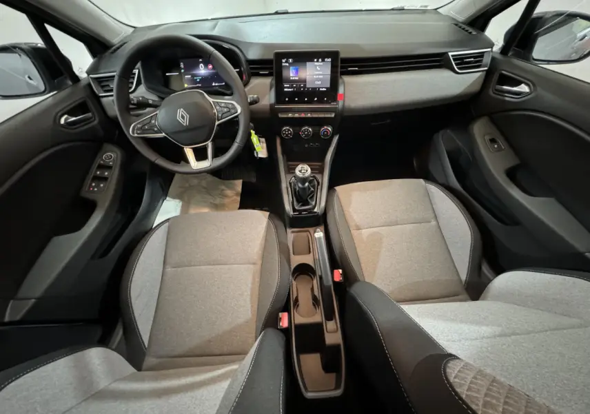 Vue intérieure avant de la Renault Clio gris clair 2025, avec volant multifonctions, écran tactile central et sièges tissu bicolore.