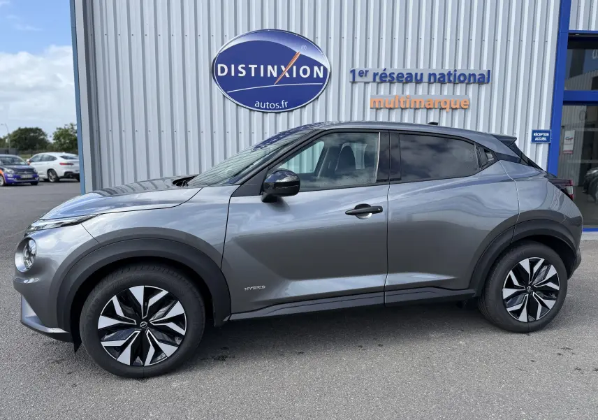 Vue de profil côté gauche du Nissan Juke Hybrid gris squale métal avec jantes au design bicolore, stationné devant un bâtiment Distinxion.