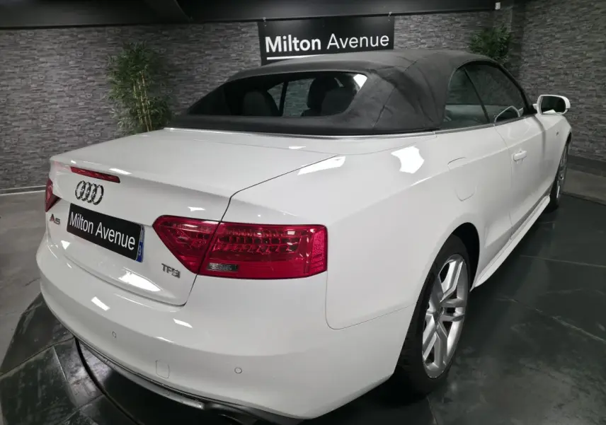 Vue 3/4 arrière droite d'un cabriolet Audi A5 blanc avec capote noire et jantes sport cinq branches doubles.