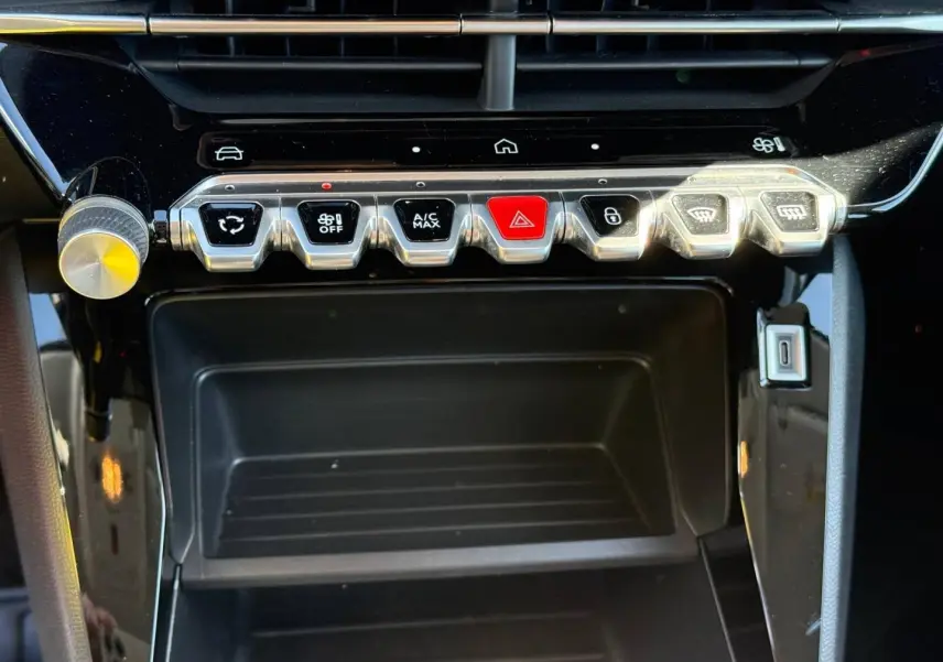 Gros plan sur la console centrale de la Peugeot 208 1.2 Hybrid 110ch Style, boutons argentés et rouge sur fond noir brillant.