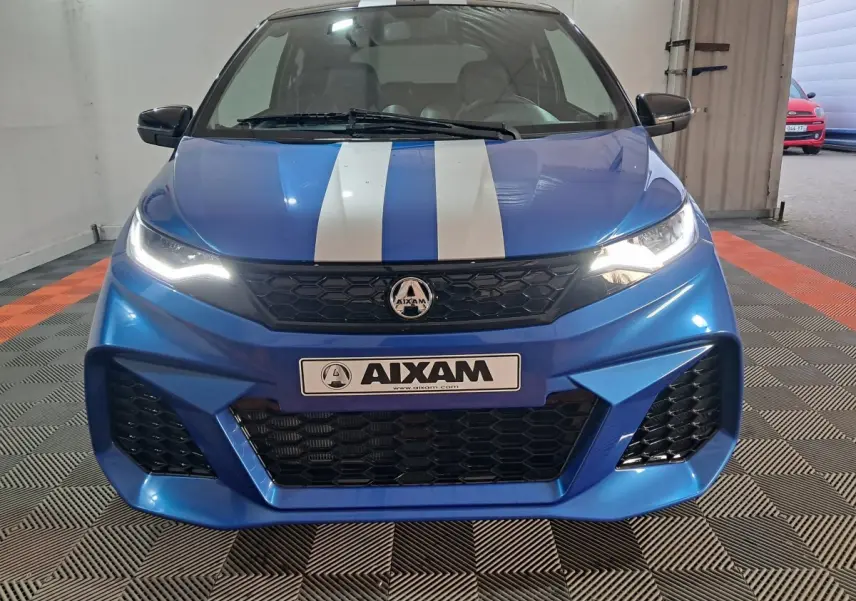 Vue frontale d'une AIXAM AMBITION 2025 bleu avec bandes blanches sur le capot et phares allumés dans un garage.