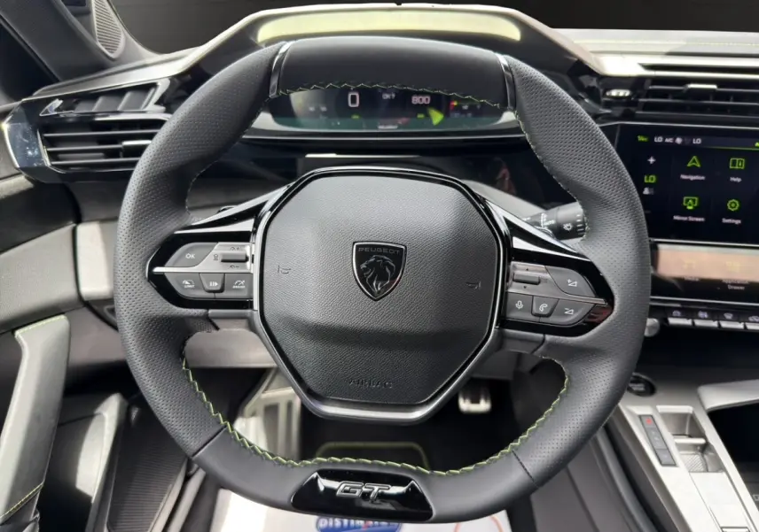 Vue rapprochée du volant cuir noir avec surpiqûres vertes et logo GT de la Peugeot 308 2026, tableau de bord digital visible.