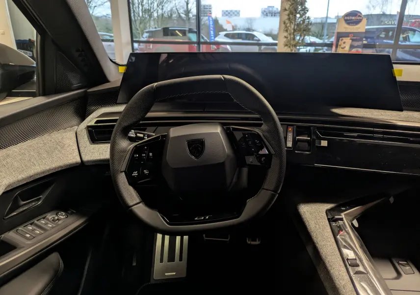 Vue intérieure centrée sur le volant GT et le tableau de bord numérique du Peugeot 3008 Hybrid gris titane, avec pédalier alu visible.