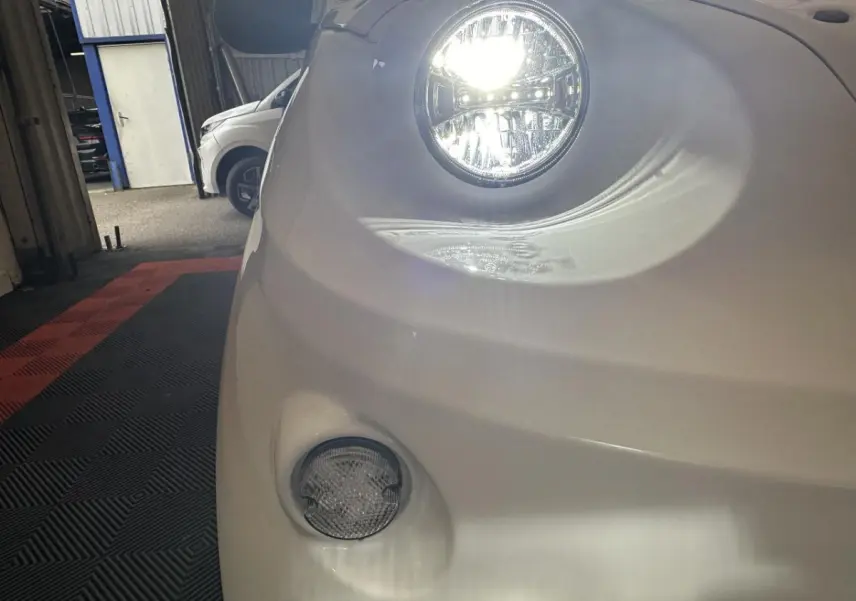 Gros plan sur le phare avant droit allumé d'une Aixam Minauto E Access blanche, avec feu antibrouillard visible.