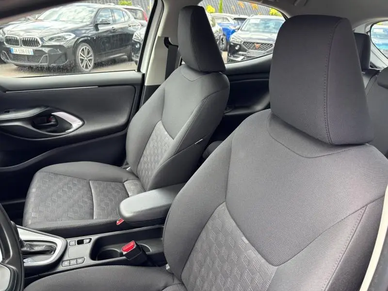Intérieur de la Toyota Yaris 116h Dynamic 5p blanc, vue rapprochée des sièges avant en tissu gris avec accoudoir central.