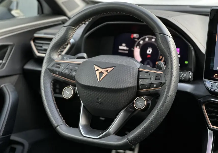 Vue rapprochée du volant cuir perforé noir avec logo CUPRA cuivré et commandes intégrées, tableau de bord numérique en arrière-plan.