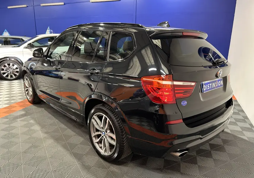 BMW X3 noir vue 3/4 arrière droit en intérieur, avec toit ouvrant et attelage visible sur le hayon.
