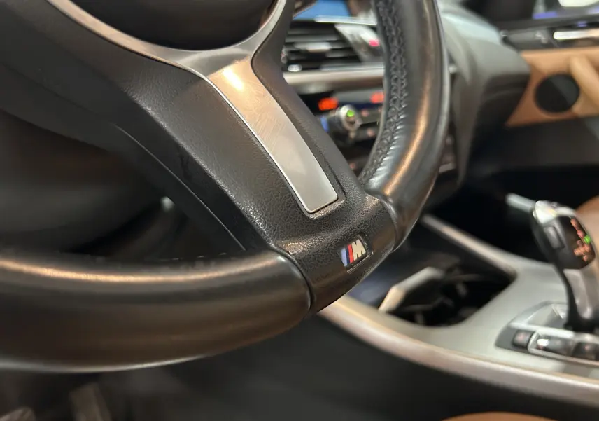 Gros plan sur le volant noir avec logo M et levier de vitesses dans l’habitacle marron d’une BMW X3 sDrive18dA 2017.