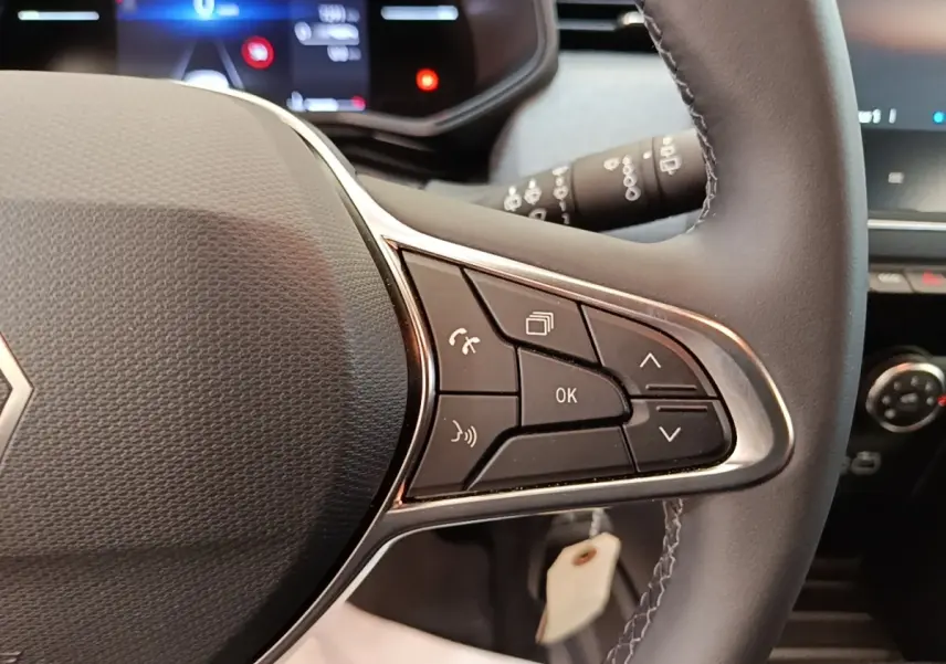 Gros plan sur le volant cuir synthétique de la Renault Clio gris clair, avec commandes multifonctions et tableau de bord numérique flou.