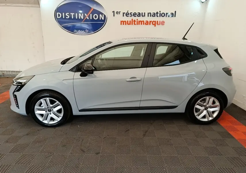 Profil côté gauche d'une Renault Clio gris clair 2025, avec jantes alliage et rétroviseurs noirs, en intérieur showroom.