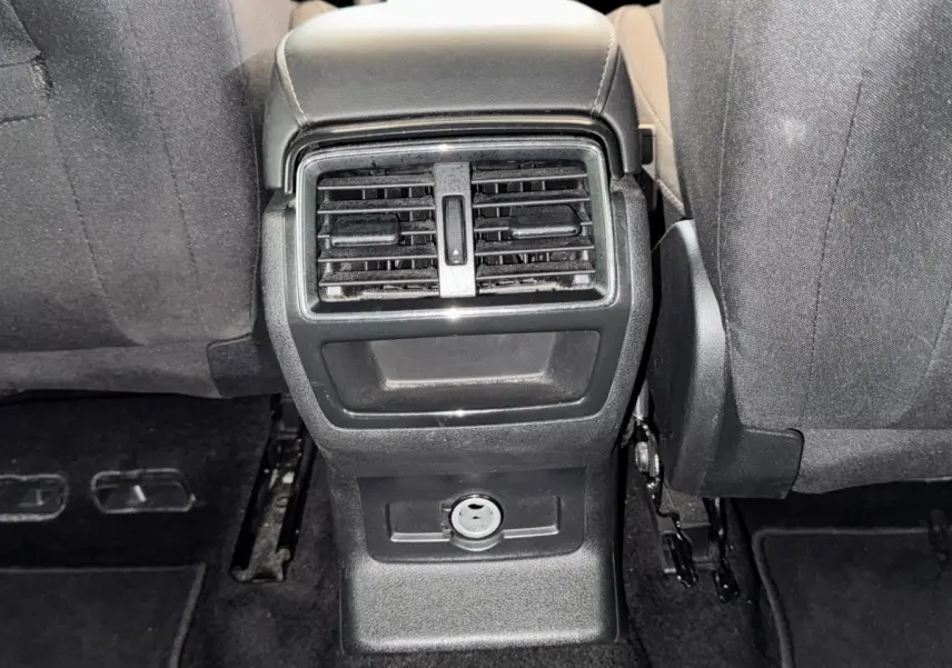 Vue rapprochée de la console centrale arrière noire avec bouches d’aération et prise 12V entre les sièges avant gris foncé du Skoda Karoq blanc.