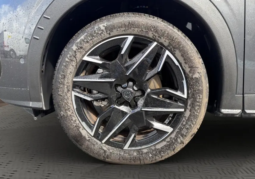 Gros plan sur la roue avant gauche d’un Peugeot 5008 gris foncé avec jante alliage noire et argentée, légèrement boueuse.