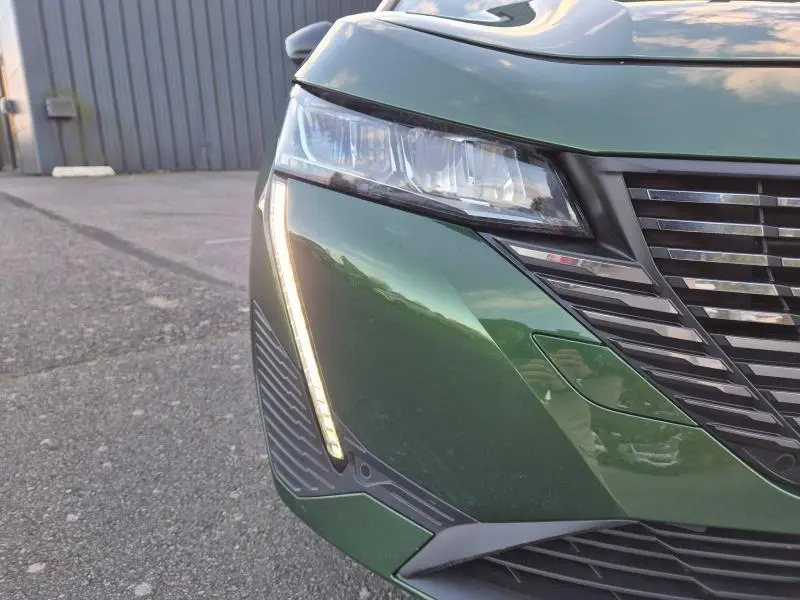 Gros plan sur le phare avant gauche allumé d'une Peugeot 308 2023 vert olivine avec calandre noire brillante.