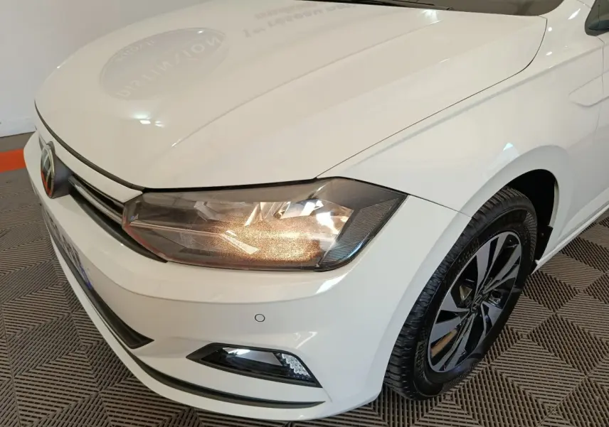 Vue 3/4 avant droit d'une Volkswagen Polo blanche avec phares allumés et jantes alliage noir et argent.